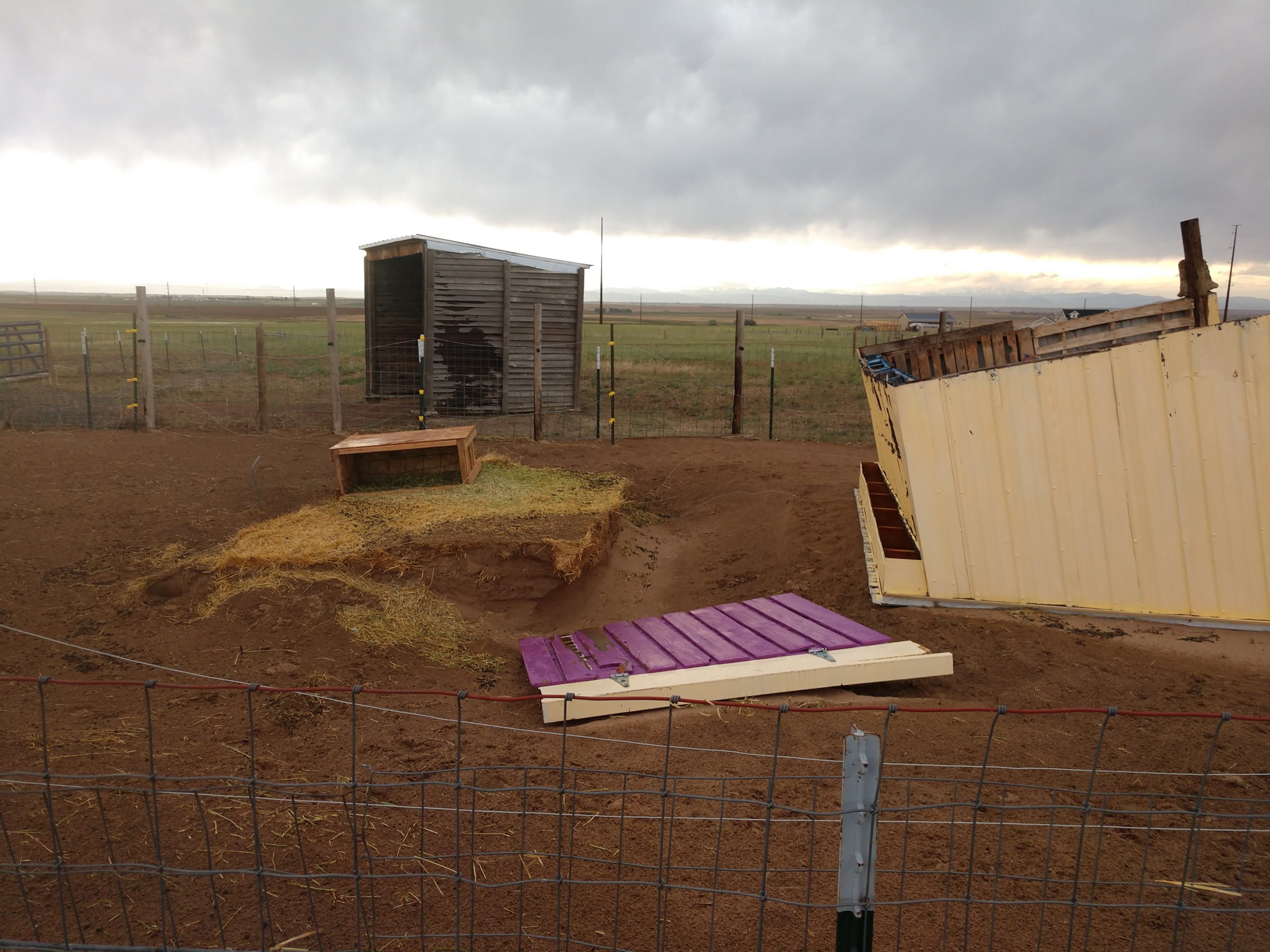 Wind-toppled shed lying on its side in a fenced dirt pen