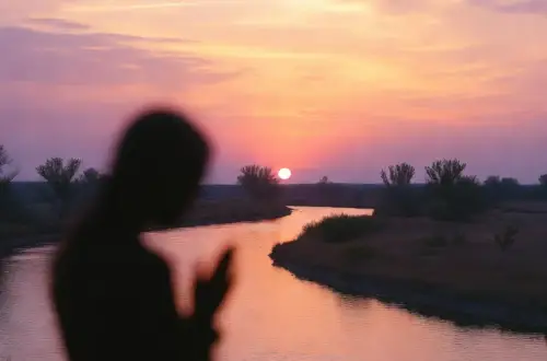 Silhouette of a woman beside a winding river at a pink and purple sunset